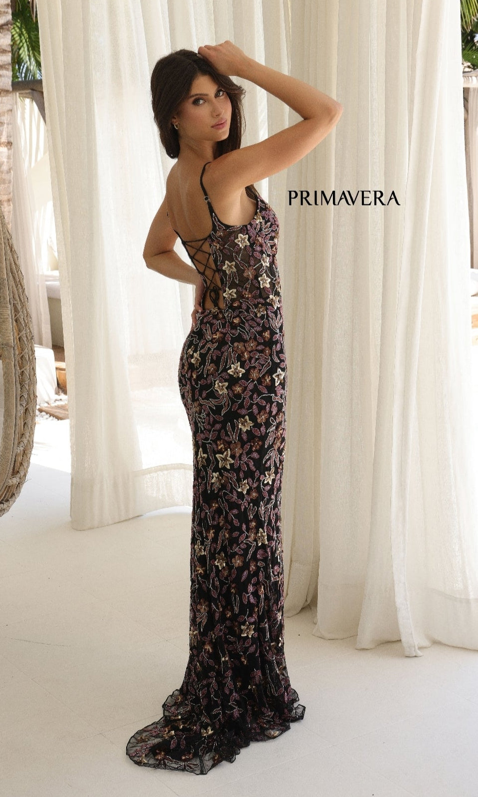 A woman stands indoors before light curtains wearing the Sequin-Floral Long Prom Dress: Primavera 4336, a floor-length, sleeveless dress with lace-up open-back and floral sequin design.